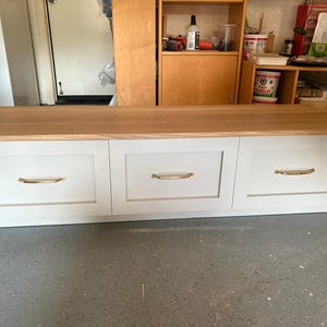 May include: A white storage bench with three drawers and a light wood top. Each drawer has a gold-colored handle. The bench is a rectangular shape and sits on a gray concrete floor.