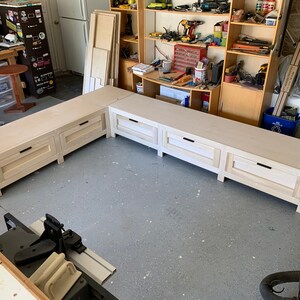 May include: A white wooden bench with drawers, featuring a corner design. The bench is unfinished and has a simple, modern style.