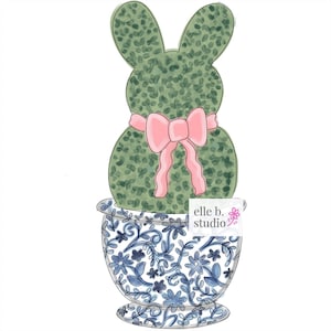 May include: A green bunny-shaped topiary plant with a pink bow tied around its neck, potted in a blue and white patterned planter with the text "elle b. studio" on the side.