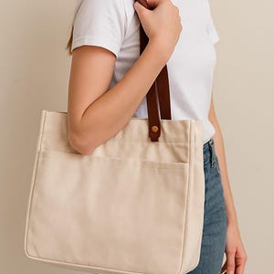 May include: Cream-colored canvas tote bag with two front pockets and brown leather handles. The bag is rectangular and has a simple, minimalist design. The handles are attached with a metal rivet.