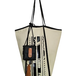 May include: A large, off-white canvas tote bag with black geometric designs. The bag has black straps and a small black pouch with a brown leather flap attached to the side. A brown tassel hangs from the bottom of the bag.