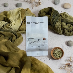 以下が含まれることがあります： 01 SOPHORA FLOWER 4 WILD DYE KITと書かれた200g用布染めキットの白いパッケージ。オリーブグリーンの染め布、緑色の毛糸玉、乾燥した種が入った小さな木製のボウル、滑らかな灰色の石に囲まれています。