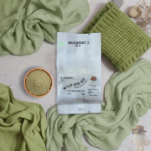 以下が含まれることがあります： Wild Dye Kit Mugwort 2と書かれた白いパッケージと小さな鳥のイラスト。オレンジ色の小さなボウルに入った緑色の粉末、緑色のニット生地、そして2枚の薄緑色のしわ加工された布が添えられています。