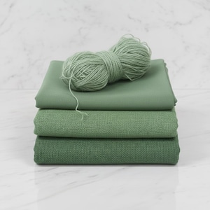 May include: A stack of three green fabric swatches with varying textures, topped with a ball of matching yarn. The fabrics range in shades from light to dark green, creating a layered effect. The yarn is a soft, muted green.