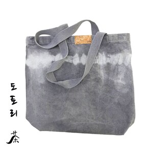 May include: A gray canvas tote bag with a tie-dye pattern in white. The bag has a cork label and long gray canvas straps.