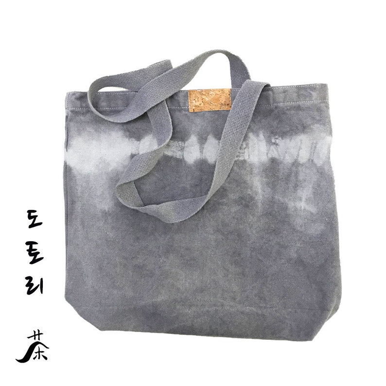 May include: A gray canvas tote bag with a tie-dye pattern in white. The bag has a cork label and long gray canvas straps.
