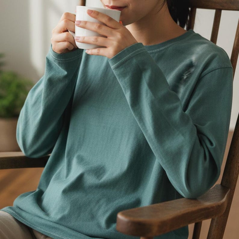 May include: A long-sleeved, teal-colored shirt. The shirt is made of a soft material and has a relaxed fit. The person is holding a white mug. The shirt is part of a casual wear collection.