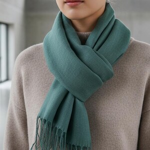 May include: A teal scarf with fringe detail is draped around a person's neck. The scarf is made of a soft material and is a solid color. The person is wearing a beige sweater.
