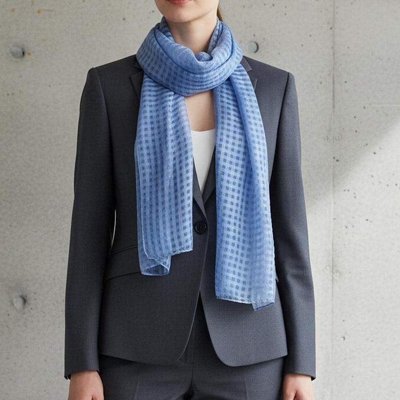 May include: A light blue scarf with a checkered pattern is draped over a dark gray blazer. The scarf is made of a lightweight fabric and adds a pop of color to the outfit. The blazer is part of a suit, suggesting a professional or formal style.
