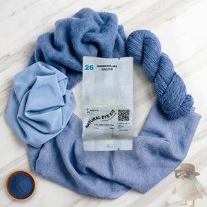 May include: A collection of blue fabric, yarn, and dye components arranged on a marble surface. The items include a skein of yarn, a folded piece of fabric, a small bowl of dye, and a package labeled "NATURAL DYE KIT" with "GARDENIA sky" text.