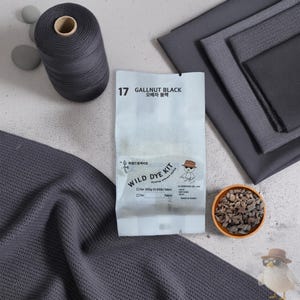 以下が含まれることがあります： Gallnut Blackの天然染料キット。白いパッケージにテキスト、ダークグレーの糸巻き、折りたたまれたダークグレーの布、乾燥した没食子が入った小さなボウル。パッケージには「17 GALLNUT BLACK」「WILD DYE KIT」「for 200g (0.45lb) fabric」と記載されています。