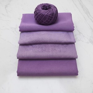 May include: A stack of four purple fabric swatches in varying shades of lavender and violet, topped with a ball of matching purple yarn. The fabrics are neatly folded, showcasing their textures and color variations.