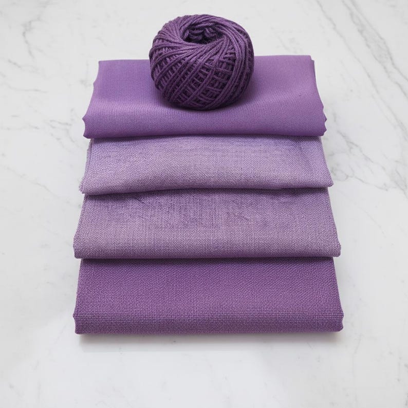 May include: A stack of four purple fabric swatches in varying shades of lavender and violet, topped with a ball of matching purple yarn. The fabrics are neatly folded, showcasing their textures and color variations.