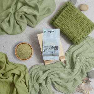 May include: An overhead shot features a natural dye kit with a knitted item and fabric swatches in shades of green. The kit includes a packet with text and a small bowl of powder. The knitted item is a green, ribbed, rectangular piece. The fabric swatches are draped.