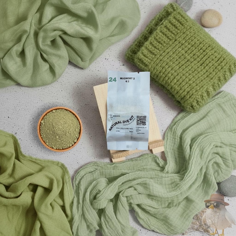 May include: An overhead shot features a natural dye kit with a knitted item and fabric swatches in shades of green. The kit includes a packet with text and a small bowl of powder. The knitted item is a green, ribbed, rectangular piece. The fabric swatches are draped.