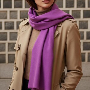 May include: A vibrant purple scarf draped over a tan trench coat. The scarf is made of a soft material and contrasts with the neutral tones of the coat and a brown turtleneck. The scarf adds a pop of color to the outfit.