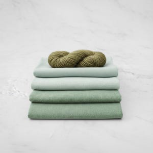May include: A stack of four folded fabric squares in shades of green, topped with a skein of olive green yarn. The fabric colors range from light mint to a deeper sage green, creating a layered effect. The yarn is thick and textured.