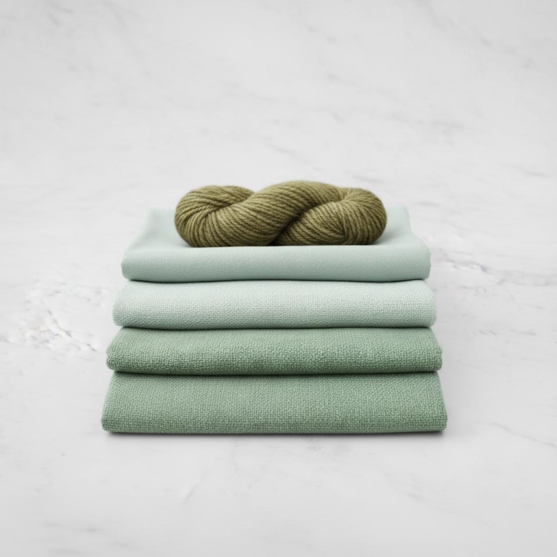 May include: A stack of four folded fabric squares in shades of green, topped with a skein of olive green yarn. The fabric colors range from light mint to a deeper sage green, creating a layered effect. The yarn is thick and textured.