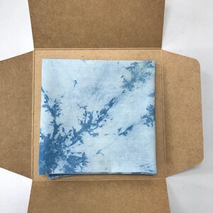 May include: A square, light blue and white tie-dye fabric is presented inside an open, brown cardboard box. The fabric has a unique pattern of blue dye that resembles tree branches.