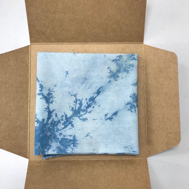 May include: A square, light blue and white tie-dye fabric is presented inside an open, brown cardboard box. The fabric has a unique pattern of blue dye that resembles tree branches.