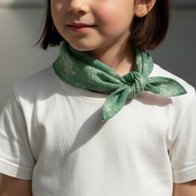 May include: A child wearing a white t-shirt and a green bandana tied around the neck. The bandana has a light green and white pattern. The bandana is knotted in the front.
