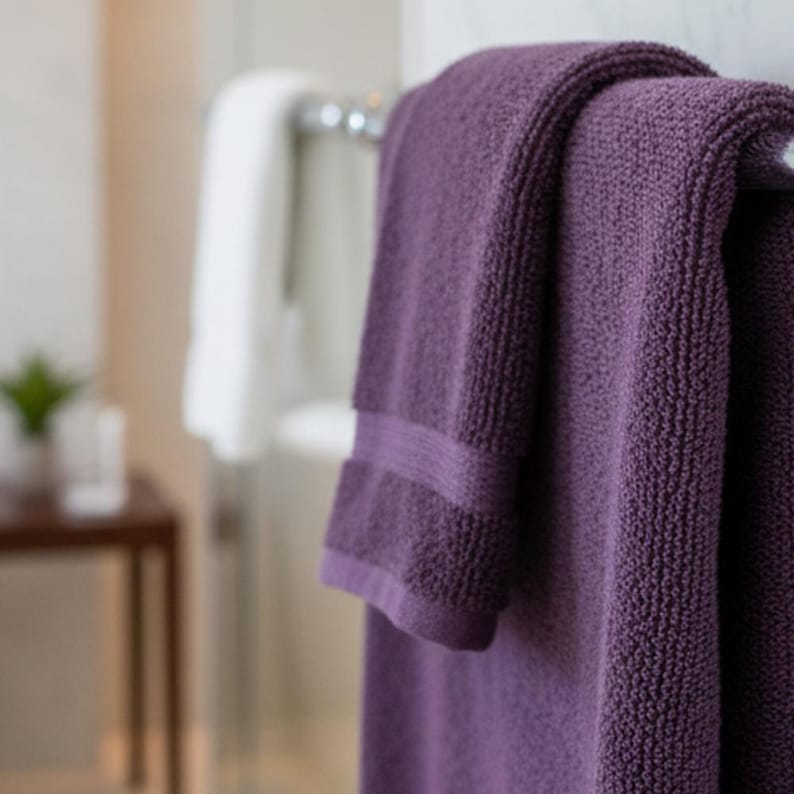 May include: A close-up of a plush, purple towel hanging on a chrome towel rack. The towel has a textured surface and a band detail near the edge. A white towel is visible in the background, along with a small table and a plant.