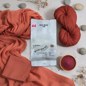 以下が含まれることがあります： 「44 RED MUD」と記されたワイルドダイキットのパッケージ。テラコッタ色の毛糸玉、折りたたまれたテラコッタ色の布、赤い染料パウダーが入った小さな木製のボウルが添えられています。このキットは200gの布地用です。