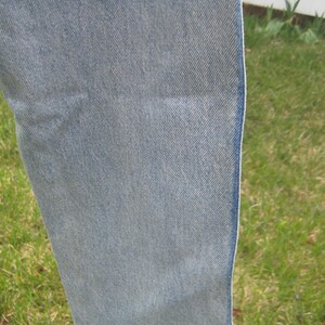 Op de afbeelding: Close-up van lichtblauwe denimstof. De denim heeft een getextureerde uitstraling met zichtbare diagonale weeflijnen. De rand van de stof is zichtbaar en toont een schone, afgewerkte naad. De achtergrond is wazig, met groen gras en een witte omheining.