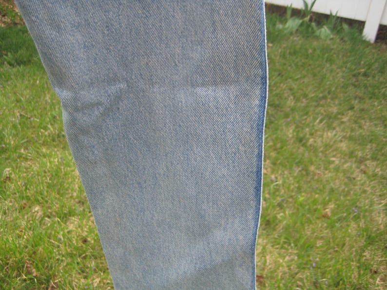 Op de afbeelding: Close-up van lichtblauwe denimstof. De denim heeft een getextureerde uitstraling met zichtbare diagonale weeflijnen. De rand van de stof is zichtbaar en toont een schone, afgewerkte naad. De achtergrond is wazig, met groen gras en een witte omheining.