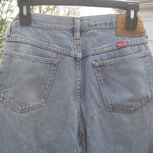 Op de afbeelding: Lichtblauwe spijkerbroek met twee achterzakken en een bruine leren patch. Het rode "Wrangler"-label is zichtbaar. De spijkerbroek hangt aan een zwarte hanger.