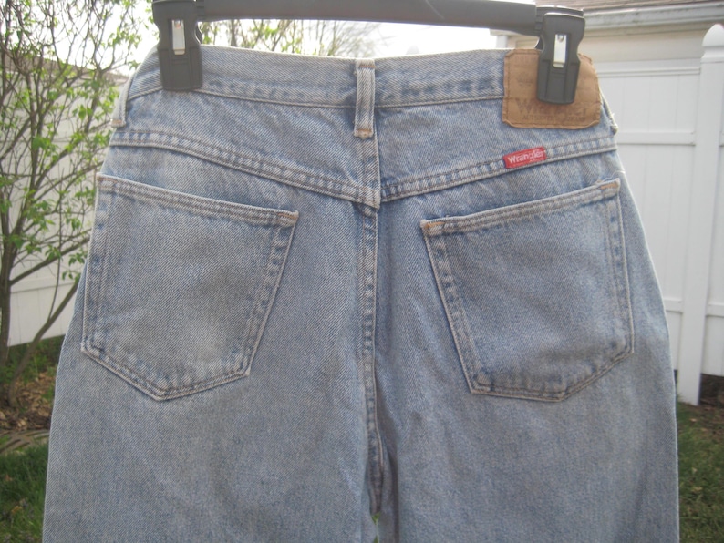 Op de afbeelding: Lichtblauwe spijkerbroek met twee achterzakken en een bruine leren patch. Het rode "Wrangler"-label is zichtbaar. De spijkerbroek hangt aan een zwarte hanger.