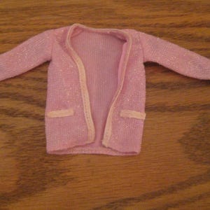 May include: A light pink long-sleeved cardigan with a subtle metallic shimmer throughout the fabric. The garment features a solid light pink trim along the open front edges, the sleeve cuffs, and two small rectangular faux pockets on the front.