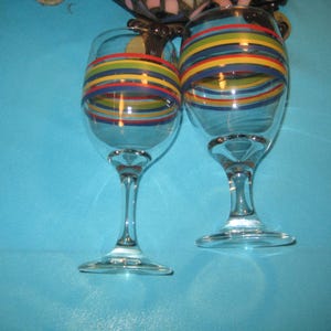 May include: Two clear glass wine glasses featuring a series of horizontal stripes in red, yellow, green, and blue encircling the upper portion of each bowl. The glasses have a standard stem and a circular base, standing upright on a surface.