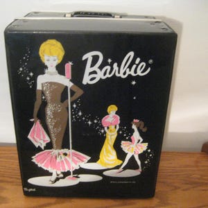 May include: A black rectangular Barbie carrying case featuring a graphic illustration of three figures in formal attire, including one standing by a microphone. The word Barbie is printed in white script at the top right, with sparkling star accents.