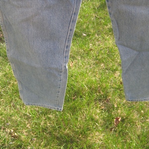 Op de afbeelding: Close-up van het onderste deel van een lichtblauwe spijkerbroek. De jeans hangt, met de benen iets uit elkaar. De stof vertoont subtiele variaties in kleur en textuur, met zichtbare naden en stiksels.