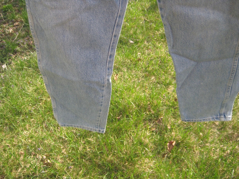 Op de afbeelding: Close-up van het onderste deel van een lichtblauwe spijkerbroek. De jeans hangt, met de benen iets uit elkaar. De stof vertoont subtiele variaties in kleur en textuur, met zichtbare naden en stiksels.