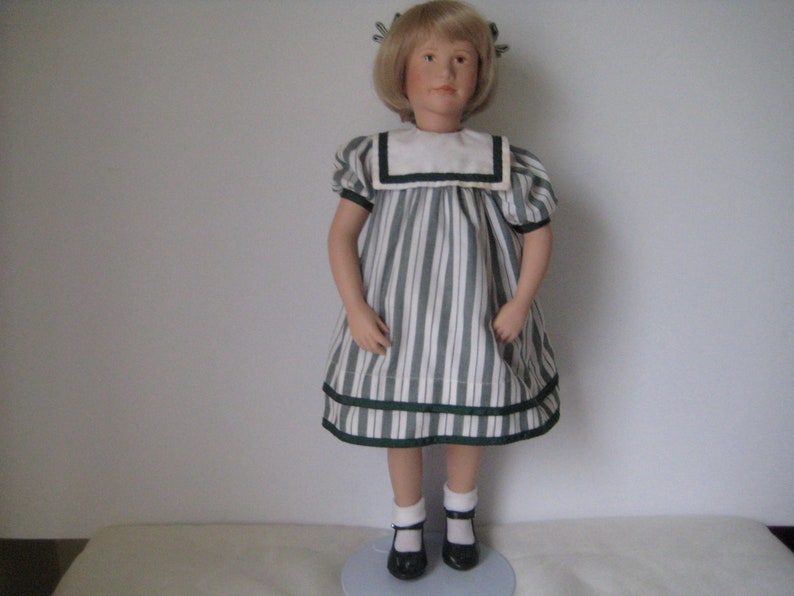 Peut inclure: Une poup&eacute;e en porcelaine portant une robe &agrave; rayures blanches et vertes avec un col marin blanc et une bordure noire. La poup&eacute;e a les cheveux blonds et les yeux bleus et porte des chaussures noires.