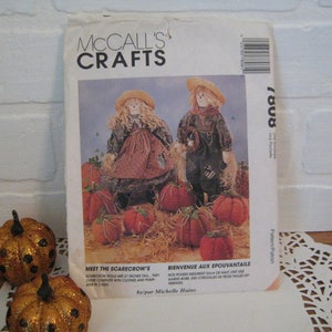 May include: A McCall's Crafts sewing pattern for making two scarecrow dolls. The pattern is titled "Meet the Scarecrow's" and "Bienvenue aux Epouvantails". The pattern shows two scarecrow dolls, one with a brown dress and the other with a black shirt and pants. The dolls are surrounded by pumpkins and straw.
