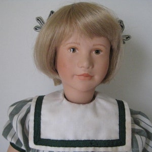 Peut inclure: Une poup&eacute;e en porcelaine aux cheveux blonds portant une robe ray&eacute;e blanche et verte avec un col blanc et une bordure verte.