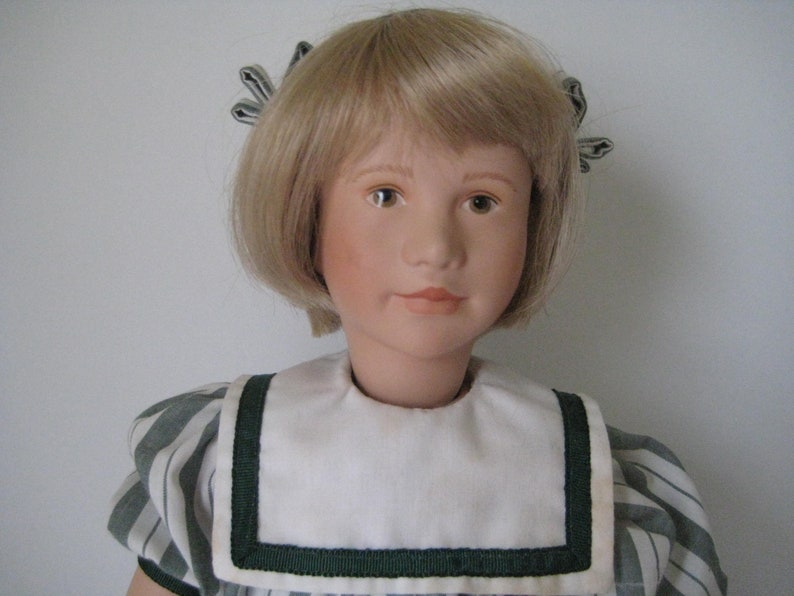 Peut inclure: Une poup&eacute;e en porcelaine aux cheveux blonds portant une robe ray&eacute;e blanche et verte avec un col blanc et une bordure verte.