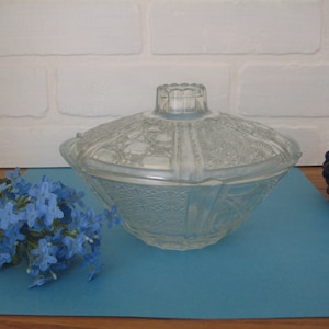 May include: A clear glass candy dish with a lid. The dish has a decorative pattern and is sitting on a blue surface.