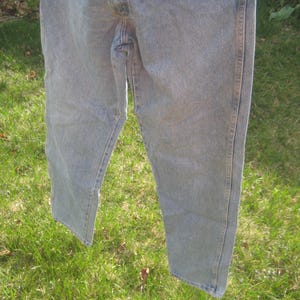 Op de afbeelding: Lichtblauwe denim jeans hangend aan een waslijn. De jeans zijn vervaagd en vertonen tekenen van slijtage. De achtergrond toont groen gras, wat een informele, alledaagse sc&egrave;ne cre&euml;ert.