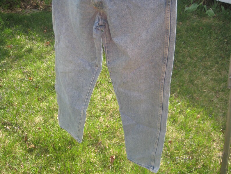 Op de afbeelding: Lichtblauwe denim jeans hangend aan een waslijn. De jeans zijn vervaagd en vertonen tekenen van slijtage. De achtergrond toont groen gras, wat een informele, alledaagse sc&egrave;ne cre&euml;ert.