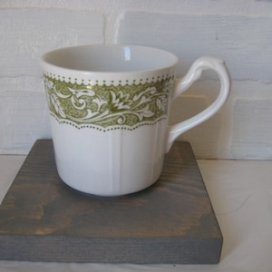 May include: A white ceramic mug with a green floral design around the top. The mug has a white handle and is sitting on a gray wooden surface.