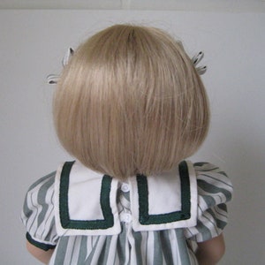 Peut inclure: Une poup&eacute;e aux cheveux blonds coiff&eacute;s en bob, portant une robe ray&eacute;e blanche et verte avec un col blanc et une bordure verte.