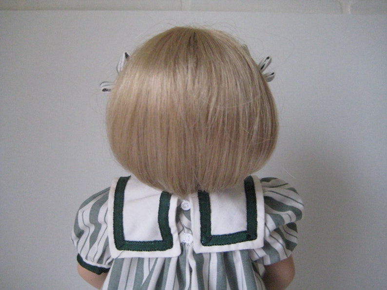 Peut inclure: Une poup&eacute;e aux cheveux blonds coiff&eacute;s en bob, portant une robe ray&eacute;e blanche et verte avec un col blanc et une bordure verte.