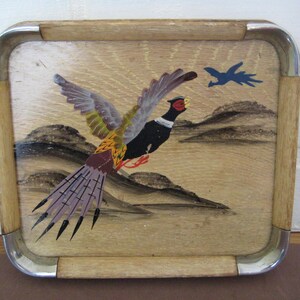 May include: A square wooden serving tray featuring a painted pheasant in flight. The pheasant has colourful feathers and is set against a light brown background with a landscape. The tray has a light wood frame with metal corner accents.