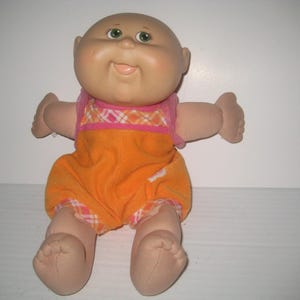 May include: A vintage Cabbage Patch Kid doll with a bald head, green eyes, and a smiling expression. The doll is wearing an orange jumpsuit with a pink and orange plaid pattern on the top and shorts. The doll's arms and legs are outstretched.
