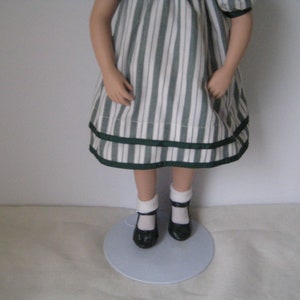 Peut inclure: Une poup&eacute;e en porcelaine portant une robe &agrave; rayures blanches et vertes avec une garniture de ruban vert. La poup&eacute;e porte des chaussettes blanches et des chaussures noires.