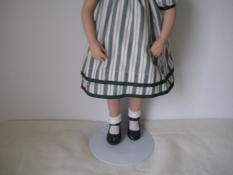 Peut inclure: Une poup&eacute;e en porcelaine portant une robe &agrave; rayures blanches et vertes avec une garniture de ruban vert. La poup&eacute;e porte des chaussettes blanches et des chaussures noires.